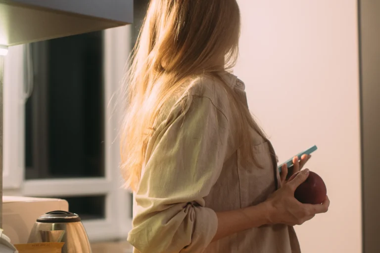 Vrouw staat in de keuken met een appel en haar gsm in de hand