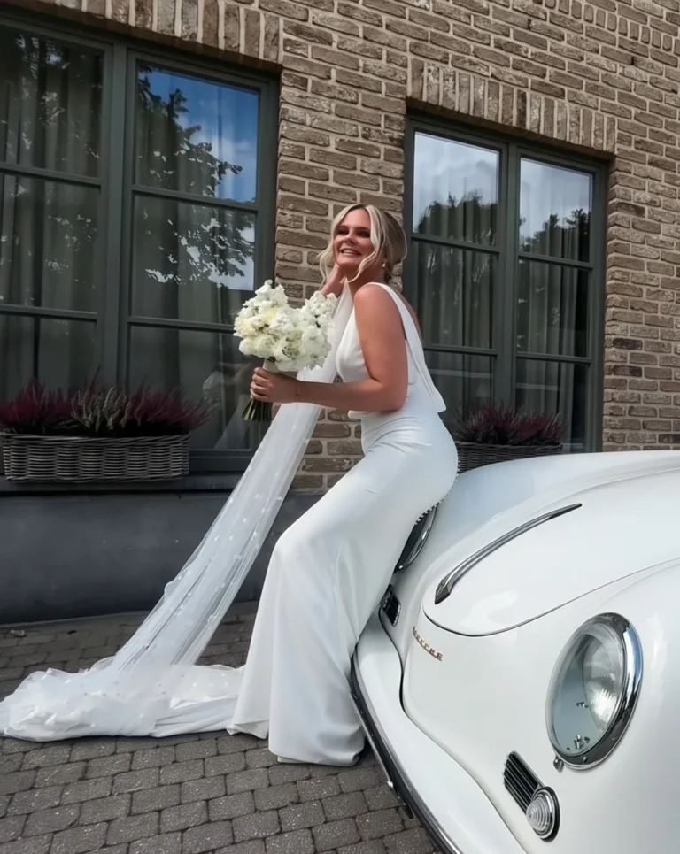 Caro, klant van Focus-On, lacht als bruid met een boeket witte bloemen, leunend op een witte oldtimer voor een bakstenen gevel
