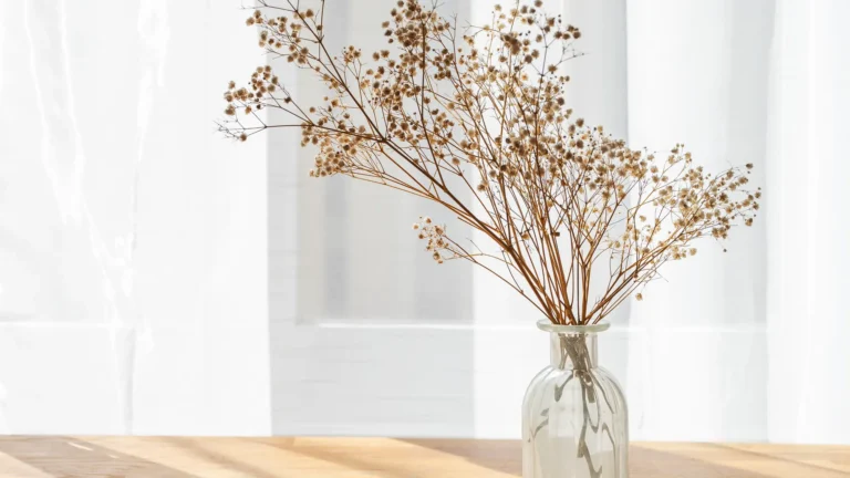Droogbloemen in een glazen vaas op een houten tafel voor een licht gordijn