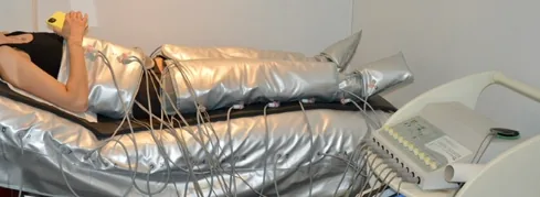 Vrouw ligt ontspannen op een behandelbed in zilveren behandelpakken met elektroden, naast het bedieningstoestel van de Firming Booster