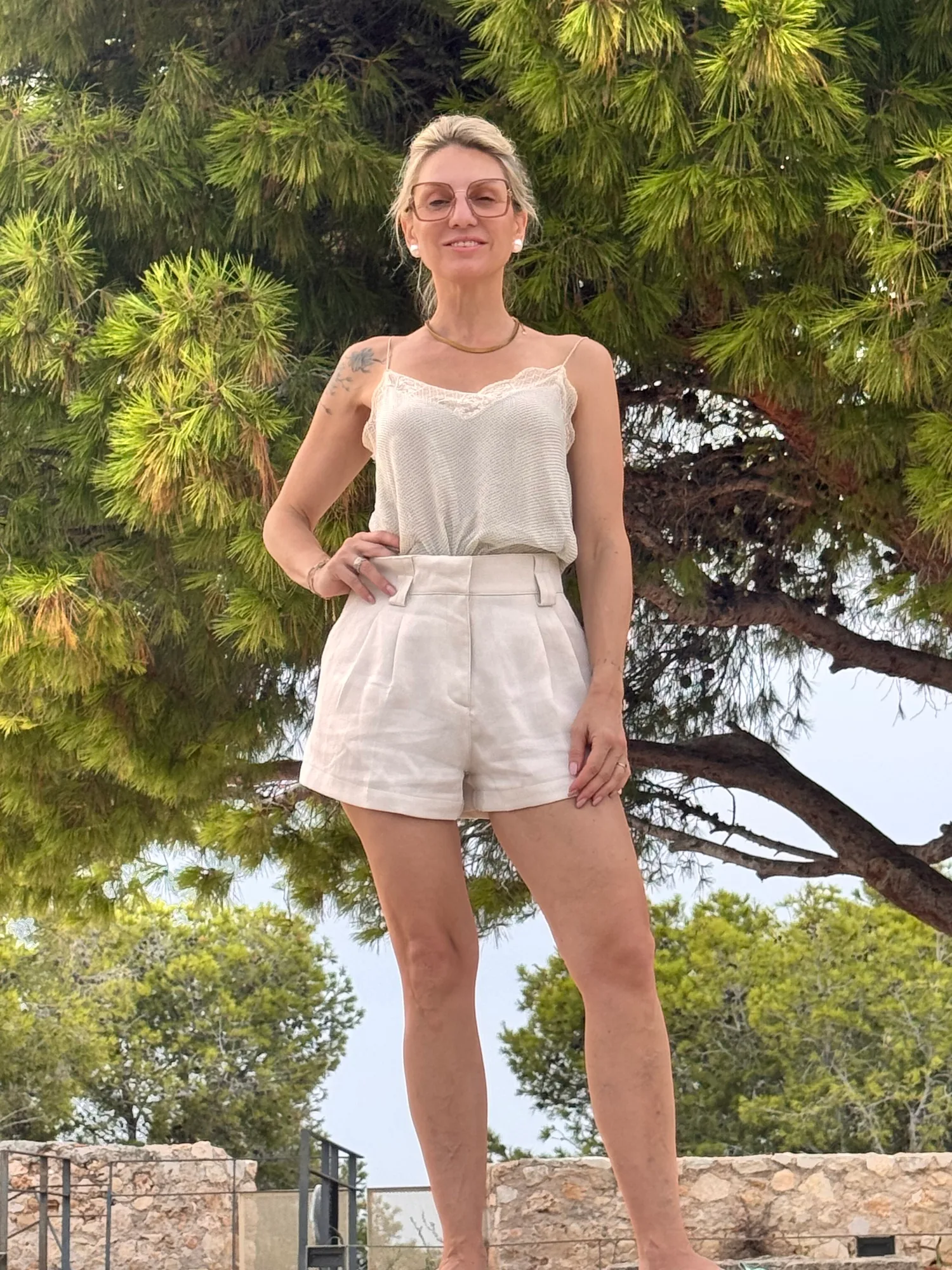 Kitty staat lachend buiten in een linnen top en short, met dennenbomen achter haar