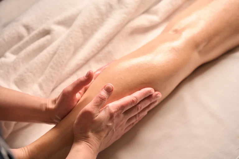 Twee handen masseren met zachte druk een onderbeen op een crèmekleurige handdoek tijdens een lymfedrainage
