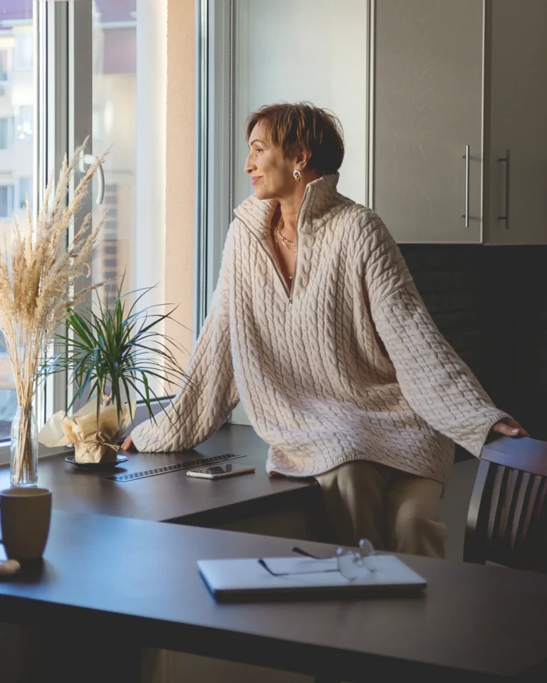 Vrouw in een crème trui leunt glimlachend tegen de werktafel en kijkt door het raam, naast droogbloemen en een laptop