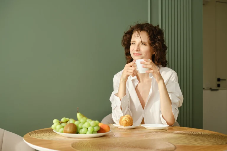 Vrouw geniet van een kop thee aan een ontbijttafel met fruit voor een saliegroene muur