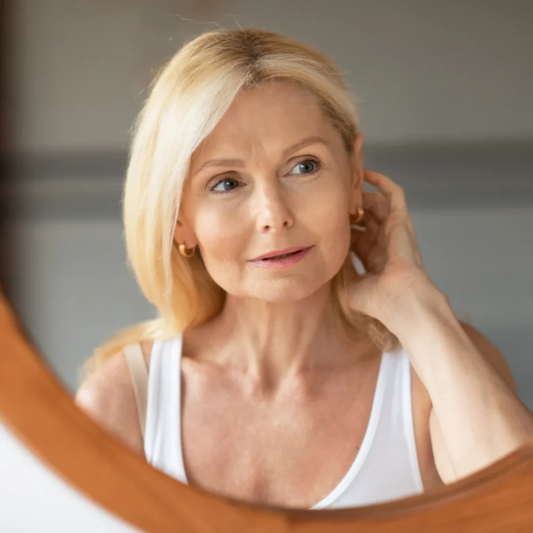 Vrouw met blond haar kijkt rustig en met een kleine glimlach naar zichzelf in een ronde spiegel met houten rand