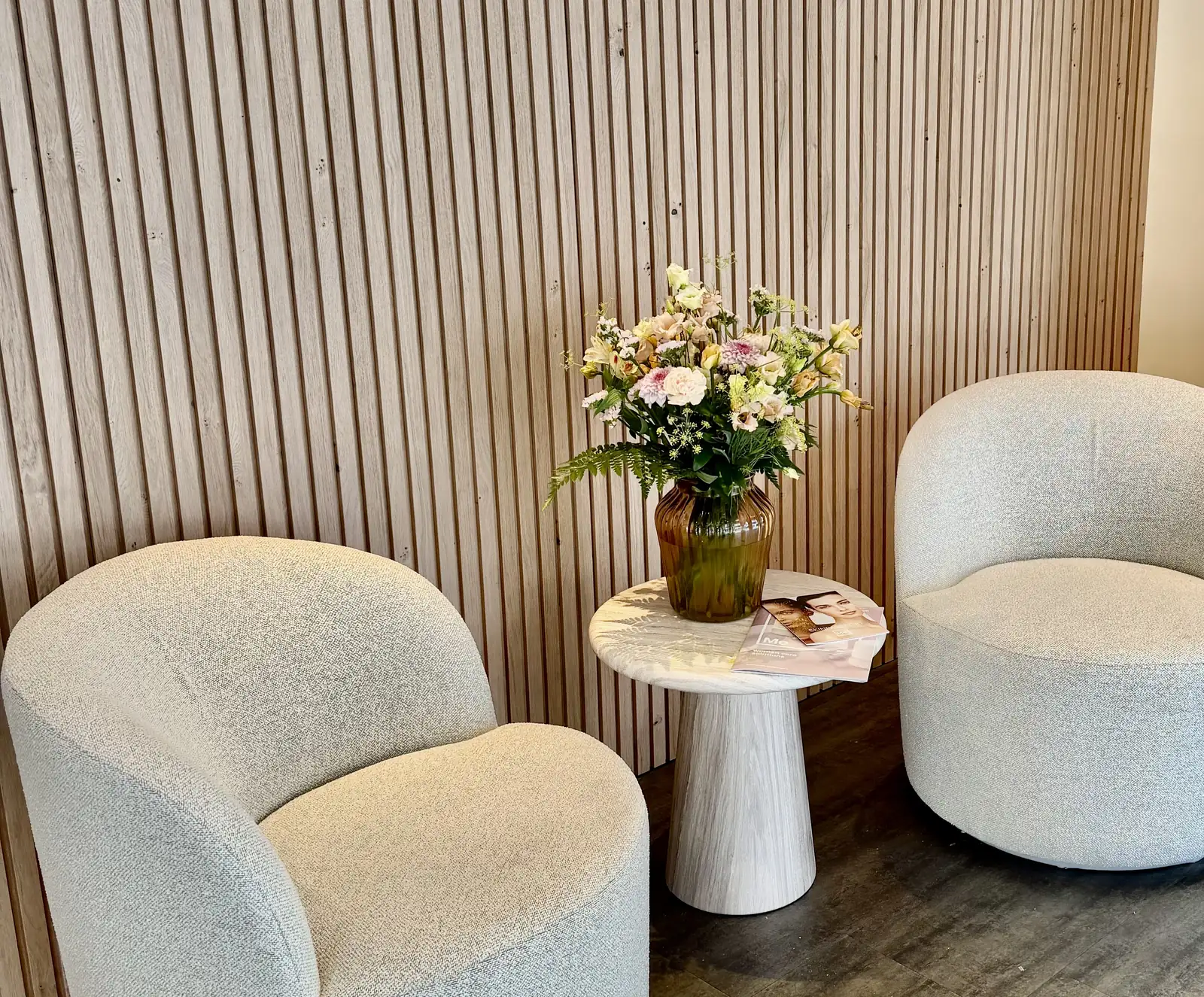 Twee witte bouclé fauteuils en een bijzettafel met een boeket voor een houten latjeswand in de wachtruimte van Focus-On