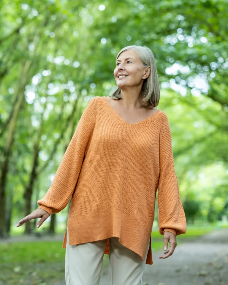 Vrouw met grijs haar en een oranje trui wandelt glimlachend over een bomenlaan in een groen park