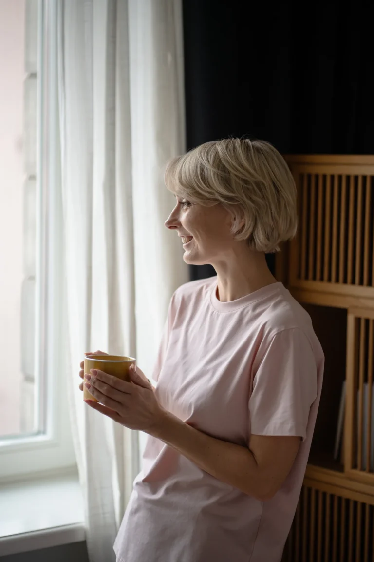 Vrouw met kort blond haar houdt een warme drank vast en kijkt glimlachend door het raam