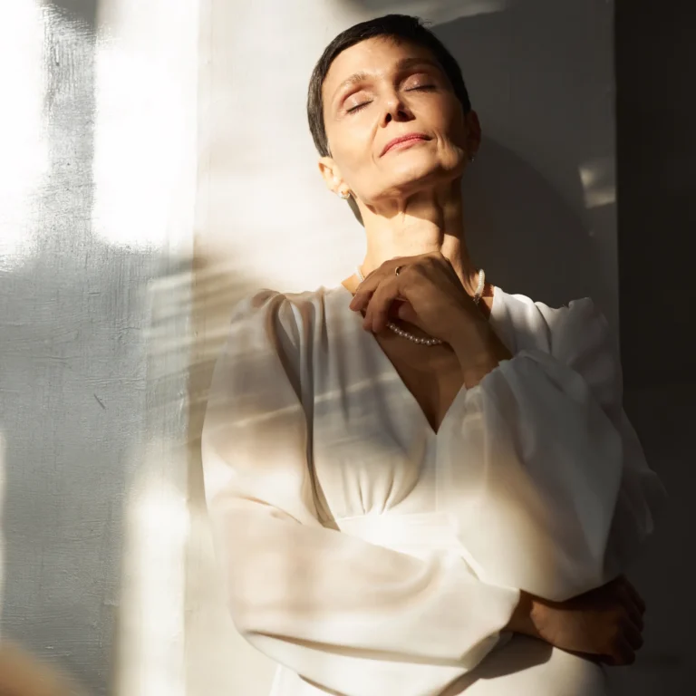 Vrouw met kort donker haar en een witte jurk staat met gesloten ogen in het zonlicht, een hand aan haar hals